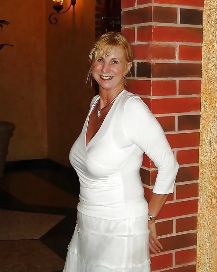 Femme mûre aux cheveux roux, souriante, portant une robe blanche, appuyée contre un mur de briques.